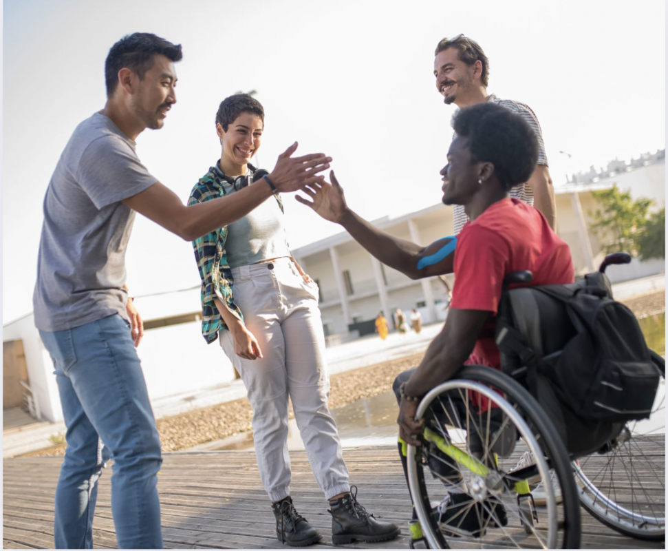 a group of people standing around a person in a wheelchair, greeting each other 